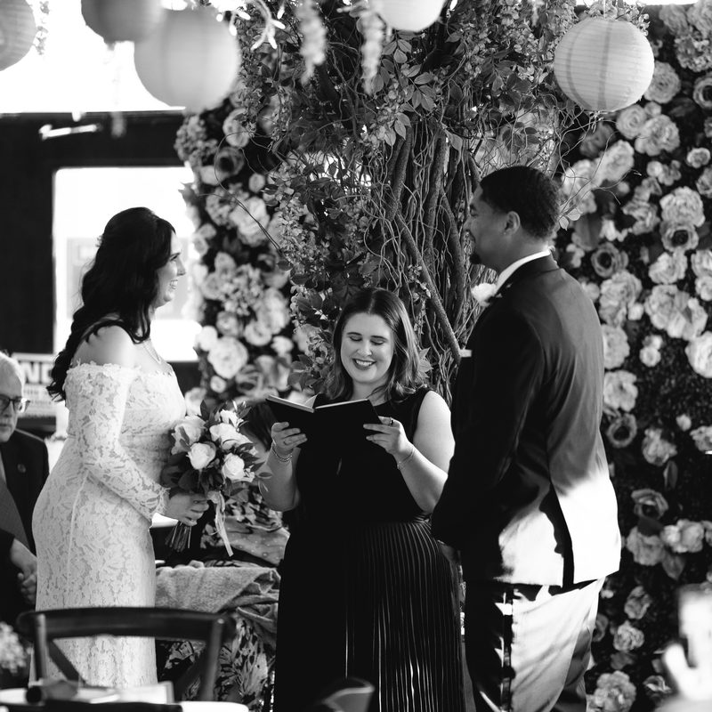 Marisa reads the ceremony to the couple beneath a floral arch hung with paper lanterns
