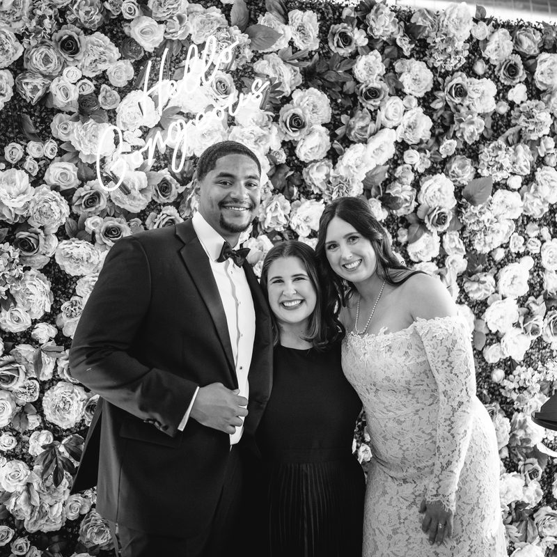 Marisa with the newlyweds in front of a white floral wall and neon sign